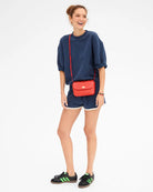A woman modeling a navy blue sweatshirt and matching shorts, accessorized with a bright red textured crossbody bag with a gold clasp, against a plain white background.