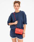 A woman modeling a navy blue sweatshirt and matching shorts, accessorized with a bright red textured crossbody bag with a gold clasp, against a plain white background.