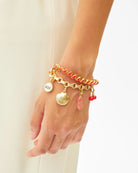 A close-up of a person's wrist and hand wearing a stack of gold charm bracelets, including a red braided bracelet, a gold chain with a shell charm, a round silver charm engraved 'OUI', and small red beads, against a light cream dress background.