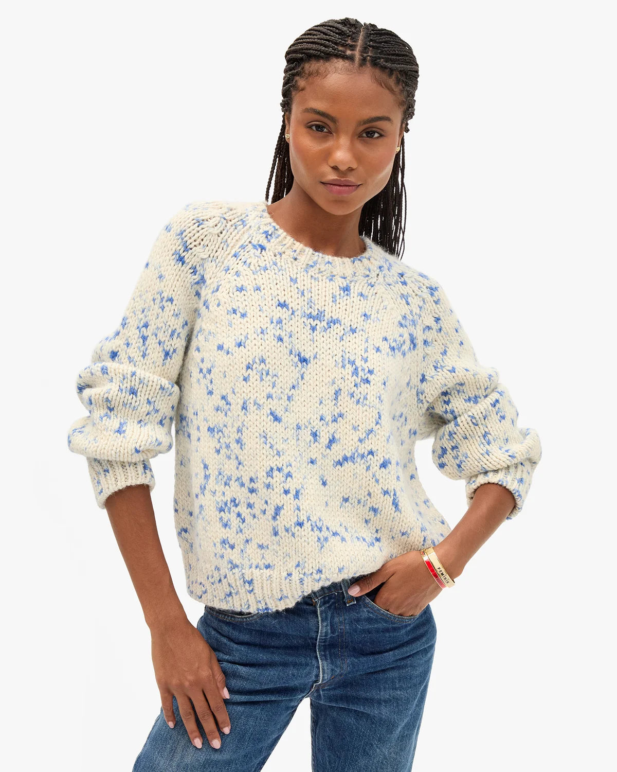 A woman modeling a cream-colored knit sweater with a blue speckled pattern, paired with blue jeans, against a plain white background.