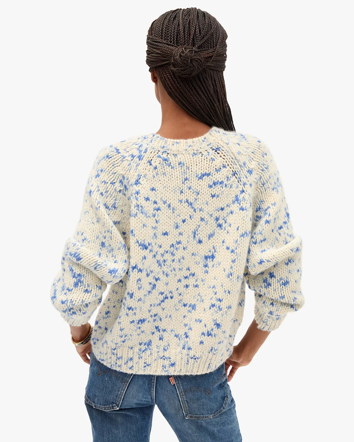 A woman viewed from behind wearing a cream-colored knit sweater with a blue floral pattern and blue jeans, set against a plain light gray background.