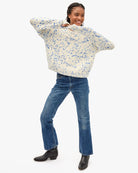 A woman modeling a cream knit sweater with a blue floral pattern, paired with blue jeans and black boots, against a plain white background.
