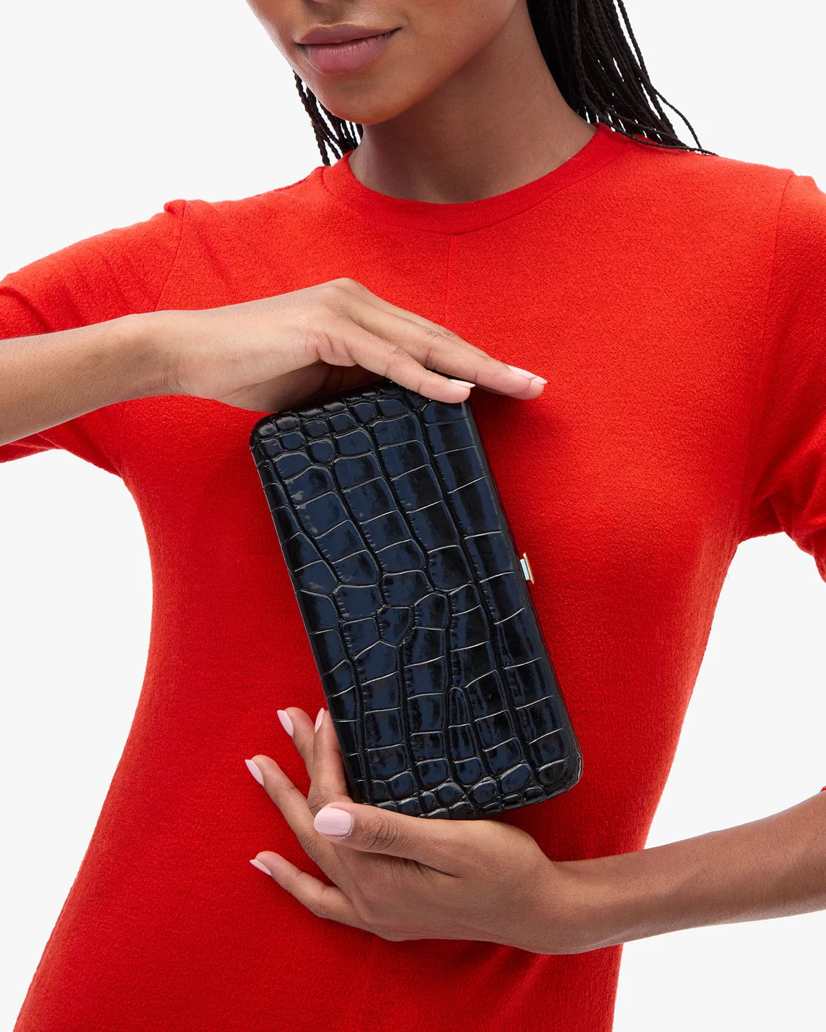 A model wearing a bright red knit top holds a black crocodile-embossed clutch bag against a clean white background.