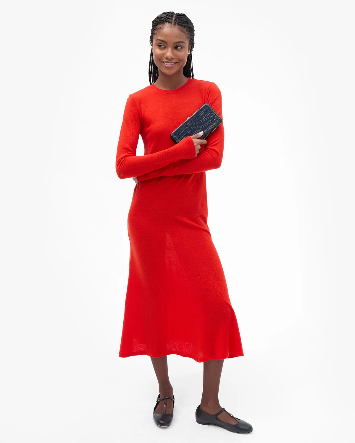 A woman modeling a bright red long-sleeve midi dress, holding a black clutch and wearing black flats, against a plain white studio background.