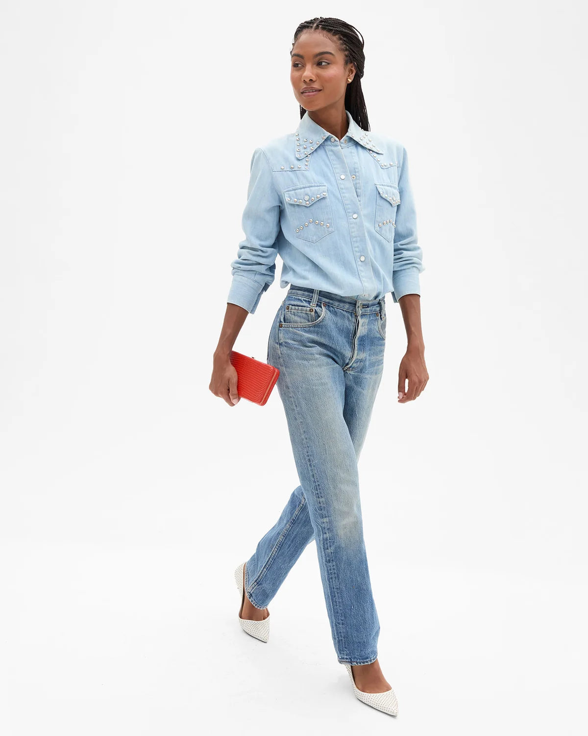 A model wearing a light blue denim western-style shirt with silver studs and matching light blue jeans, holding a small red clutch, against a plain white studio background.