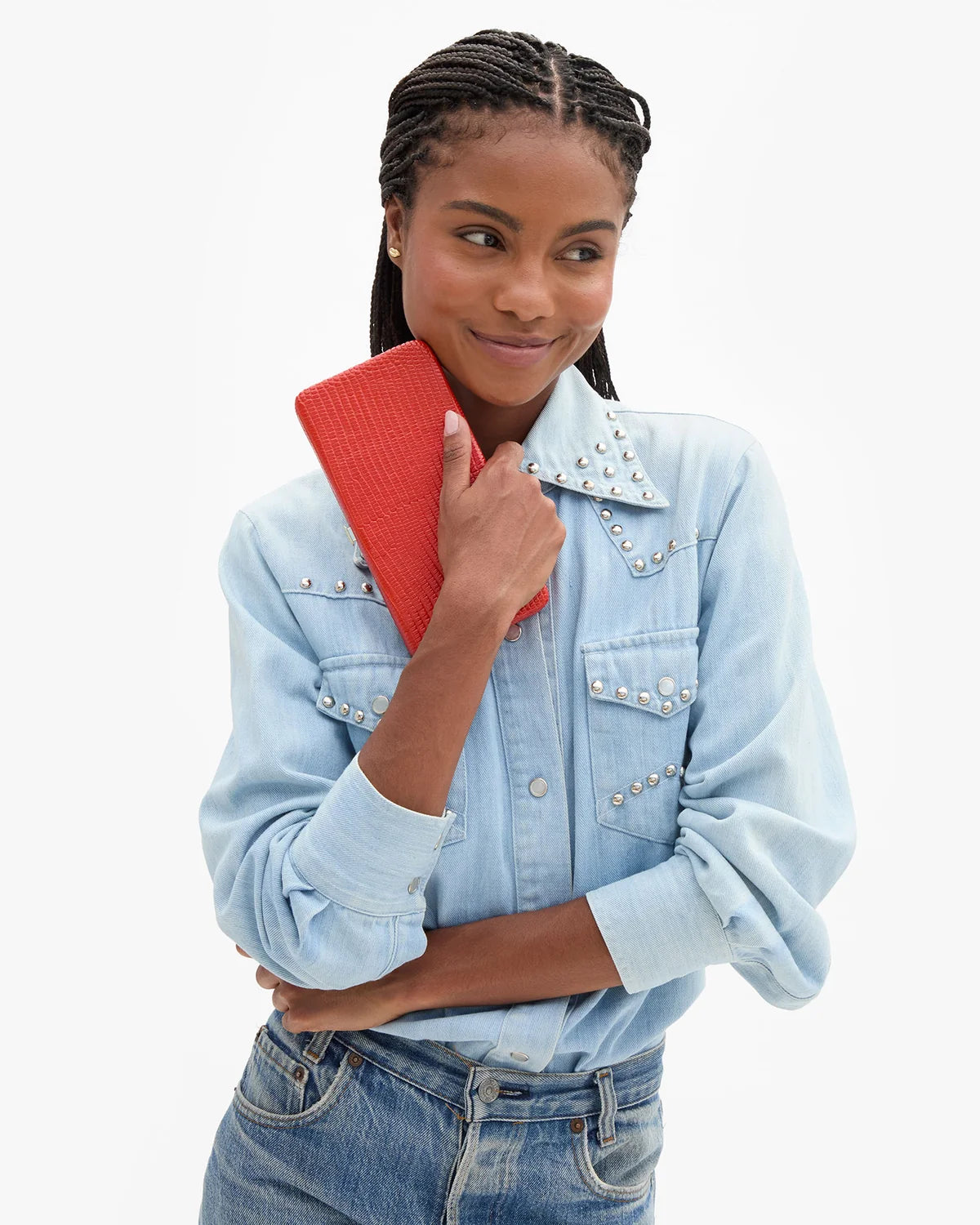 A woman modeling a light blue denim shirt with silver studs on the collar and chest, paired with blue jeans and holding a textured red clutch bag against a plain white background.