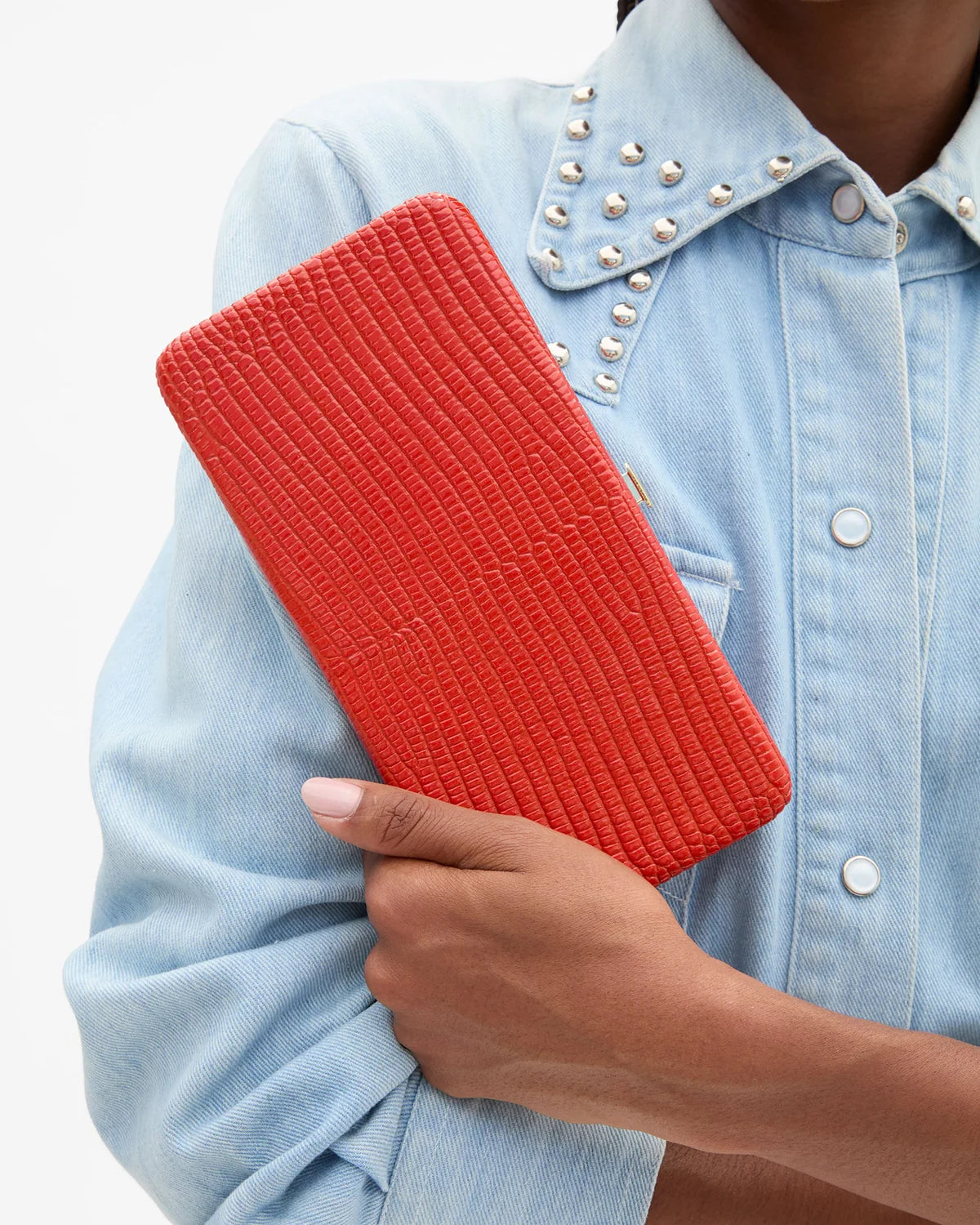 A person in a light blue denim shirt holding a bright red textured clutch purse against a clean white background.