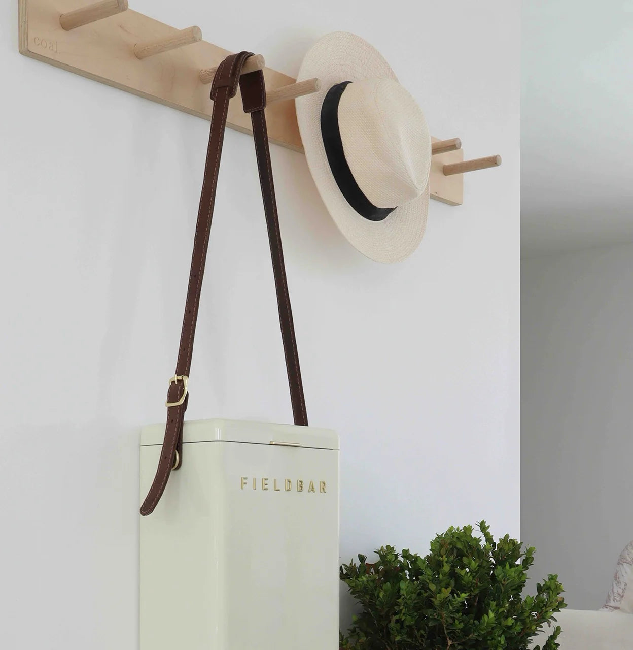 White tote bag with 'FIELDBAR' branding, straw hat, and brown strap hanging on a wooden hook against a white wall.