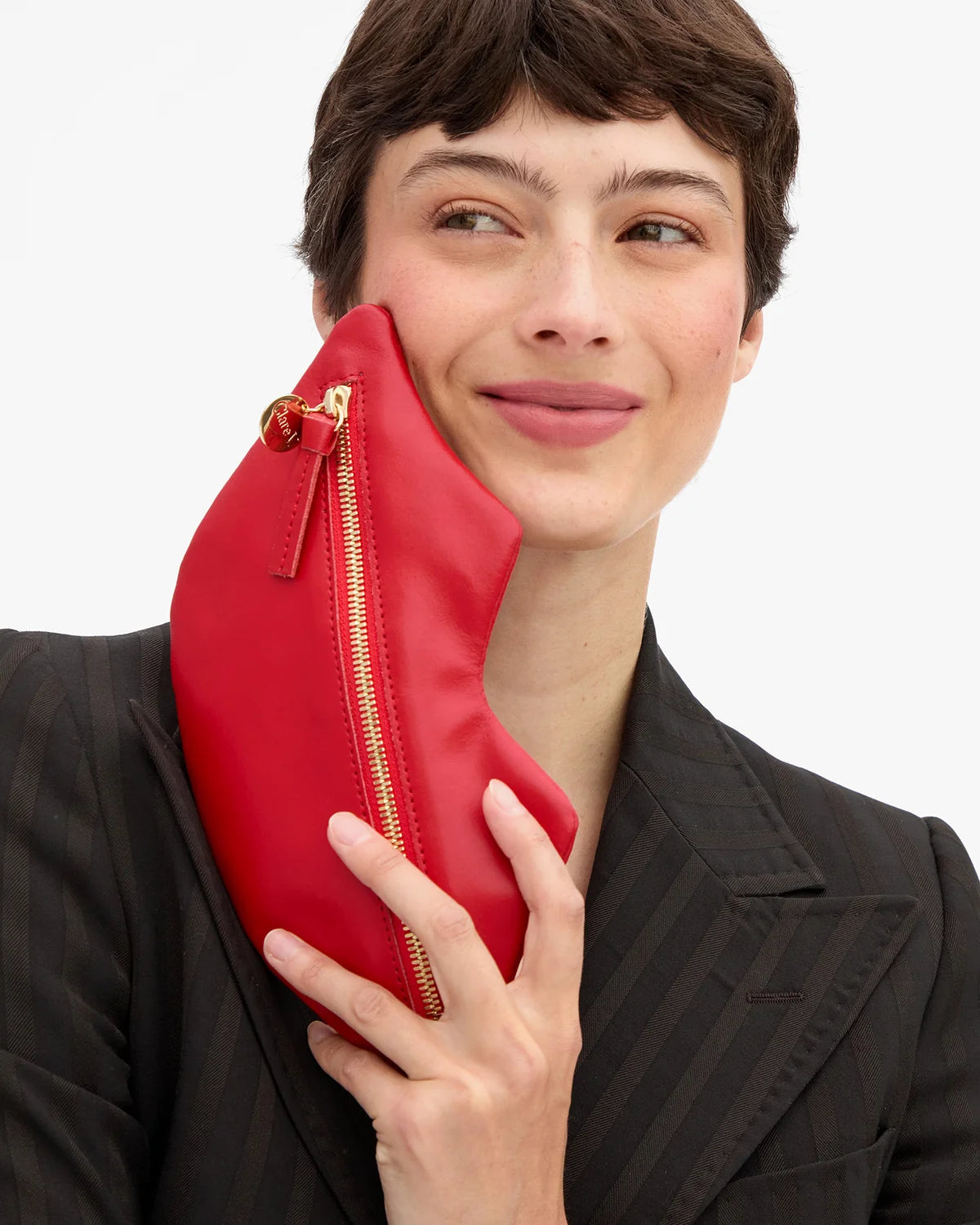 A model in a black pinstripe blazer holds a bright red leather clutch bag with a gold zipper against a clean white background.