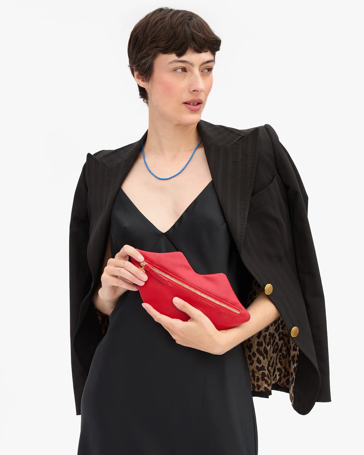A model wearing a black blazer over a black dress, holding a bright red leather clutch bag with gold hardware against a plain white background.