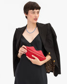 A model wearing a black blazer over a black dress, holding a bright red leather clutch bag with gold hardware against a plain white background.