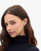 A close-up portrait of a woman with long brown hair wearing a small gold crescent moon stud earring, set against a plain light gray background.