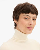 A close-up portrait of a woman with short brown hair wearing a cream ribbed turtleneck sweater and a small gold stud earring, set against a plain white background.
