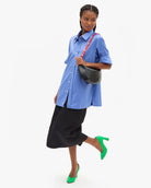 A model wearing a light blue short-sleeve button-up shirt, a black midi skirt, neon green heels, and a black crescent bag with a red strap, posed against a solid white background.
