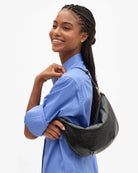 A woman with braided hair wearing a blue shirt carries a black textured leather shoulder bag against her shoulder, photographed against a clean white background.