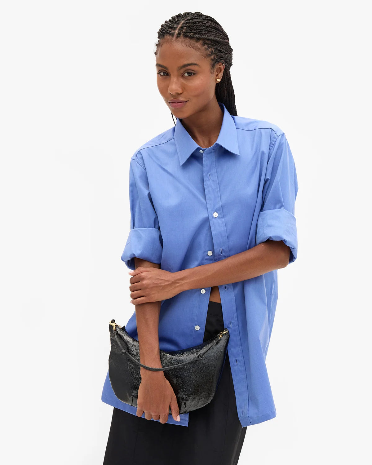 A model wearing a light blue button-down shirt with rolled-up sleeves and a black crescent-shaped shoulder bag, set against a plain white studio background.