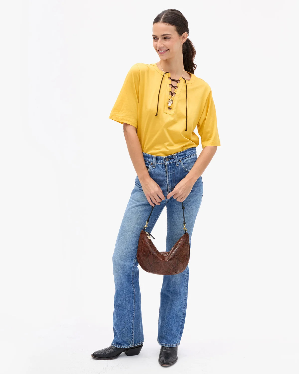 A woman modeling a bright yellow short-sleeve blouse with lace-up detailing at the neckline, paired with blue jeans and a brown crescent shoulder bag, against a plain white studio background.