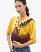 A person wearing a yellow top and blue jeans carries a brown snakeskin-patterned crescent shoulder bag with a black strap against a plain white background.