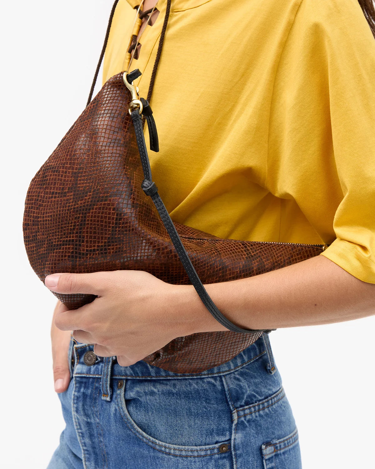 A person wearing a yellow top and blue jeans carries a brown snakeskin-patterned crescent shoulder bag with a black strap against a plain white background.