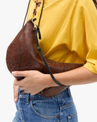 A person wearing a yellow top and blue jeans carries a brown snakeskin-patterned crescent shoulder bag with a black strap against a plain white background.
