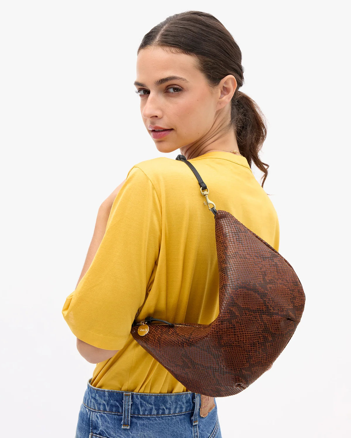 A person wearing a yellow top and blue jeans carries a brown snakeskin-patterned crescent shoulder bag with a black strap against a plain white background.