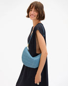 A woman wearing a black dress models a light blue woven crescent shoulder bag with a matching strap against a plain white background.