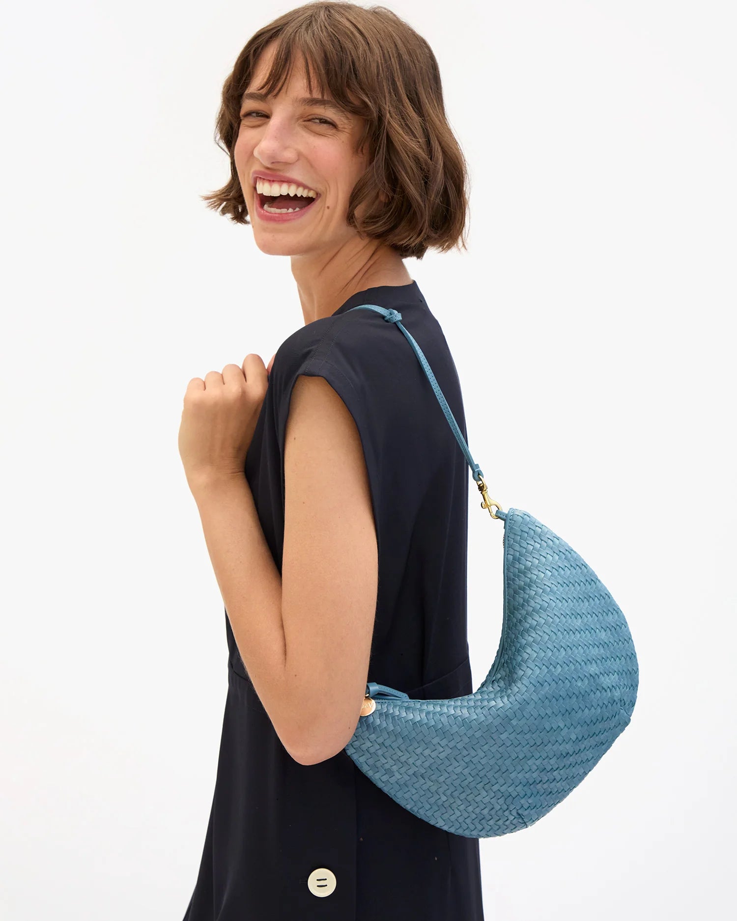 A woman with a short brown bob haircut smiling and looking back over her shoulder while wearing a blue woven crescent-shaped shoulder bag with a thin blue strap and gold-tone hardware, paired with a dark navy sleeveless top against a plain white background.
