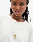 A close-up of a model wearing a delicate gold chain necklace with a multi-piece pendant featuring a white pearl, a peach-colored bead, and small green and gold details, paired with a white crewneck top.