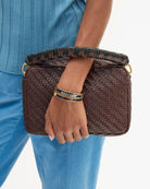 A person wearing a blue ribbed top and blue pants holds a dark brown woven clutch bag with a braided handle and gold-tone hardware. The person is also wearing a black and gold Marc Jacobs bracelet.