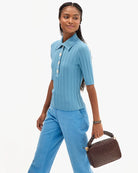 A woman modeling a light blue ribbed short-sleeve button-front top and matching blue trousers, holding a brown woven handbag against a plain light gray studio background.