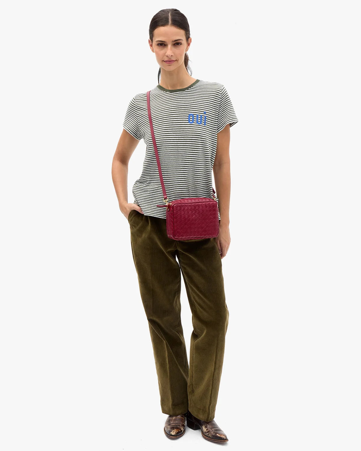 A model stands against a light gray studio background wearing a green and white striped t-shirt with a blue logo, olive green trousers, and brown leather loafers, while carrying a dark red woven crossbody bag.