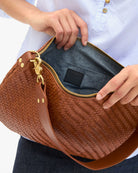 Brown woven handbag with gold zipper pull held by a person wearing a white shirt.