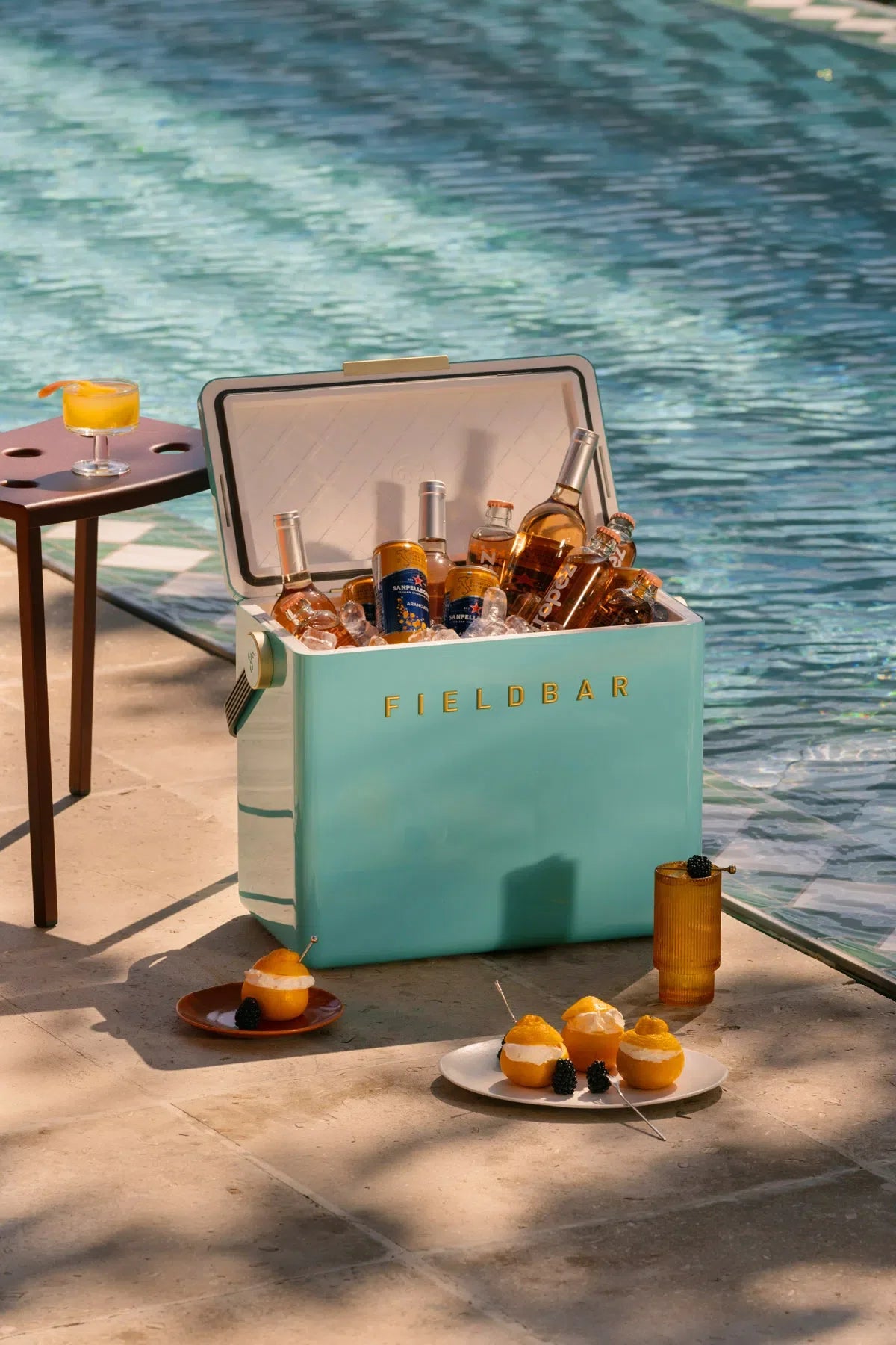 Teal cooler labeled 'FIELDBAR' filled with drinks by a poolside.