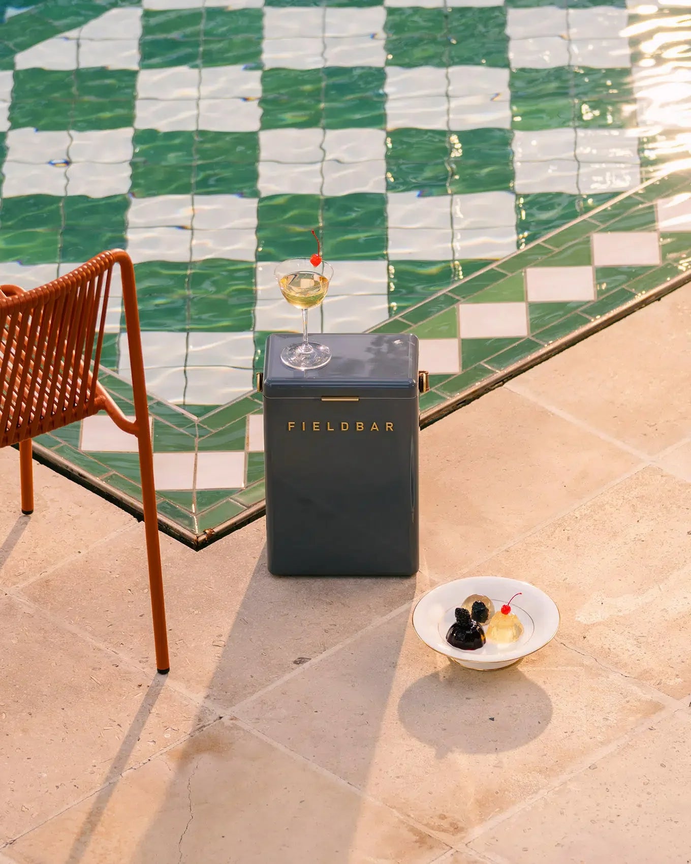 Small refrigerator labeled 'FIELDBAR' next to a chair and a small table with a glass of wine and dessert on a tiled patio.