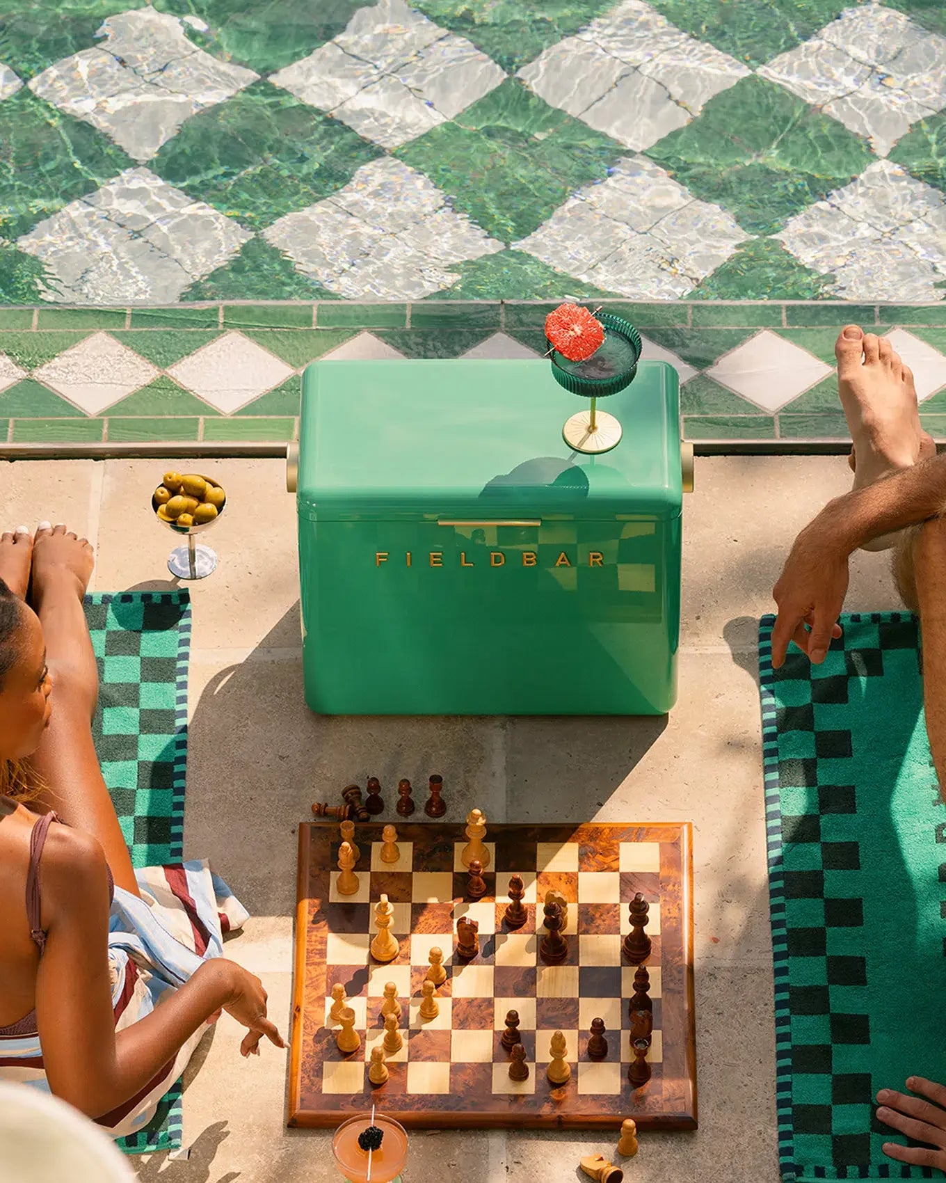 Two people playing chess outdoors with a green Fieldbar cooler in the background.
