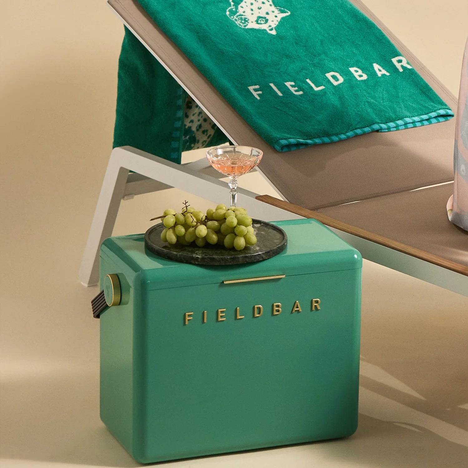 Teal 'FIELDBAR' cooler with grapes and a cocktail on a lounge chair with a green towel.