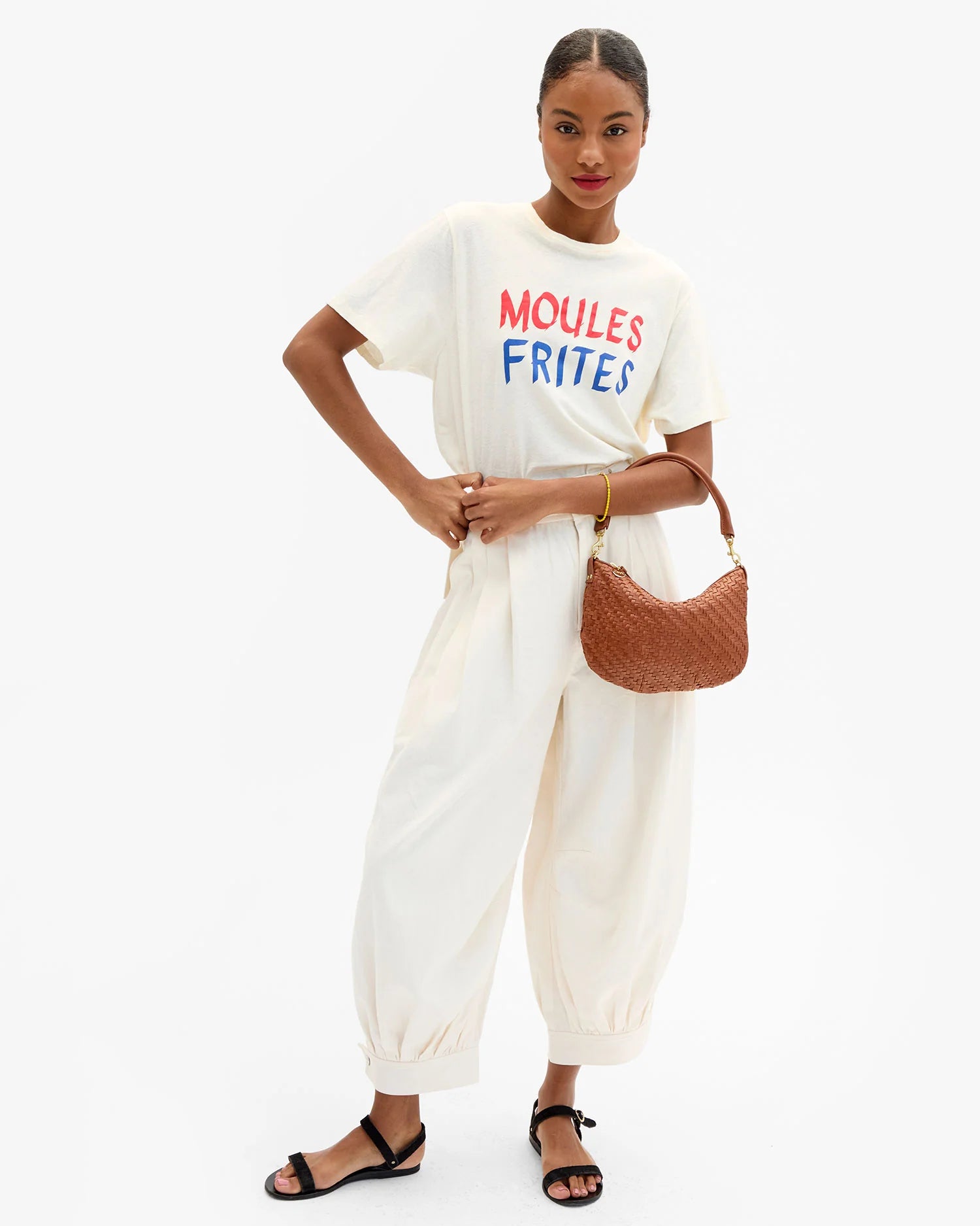 Woman wearing a white t-shirt with colorful text and white pants, holding a brown bag on a white background