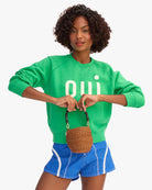 Woman wearing a green sweatshirt with 'Oui' text, holding a woven handbag on a white background