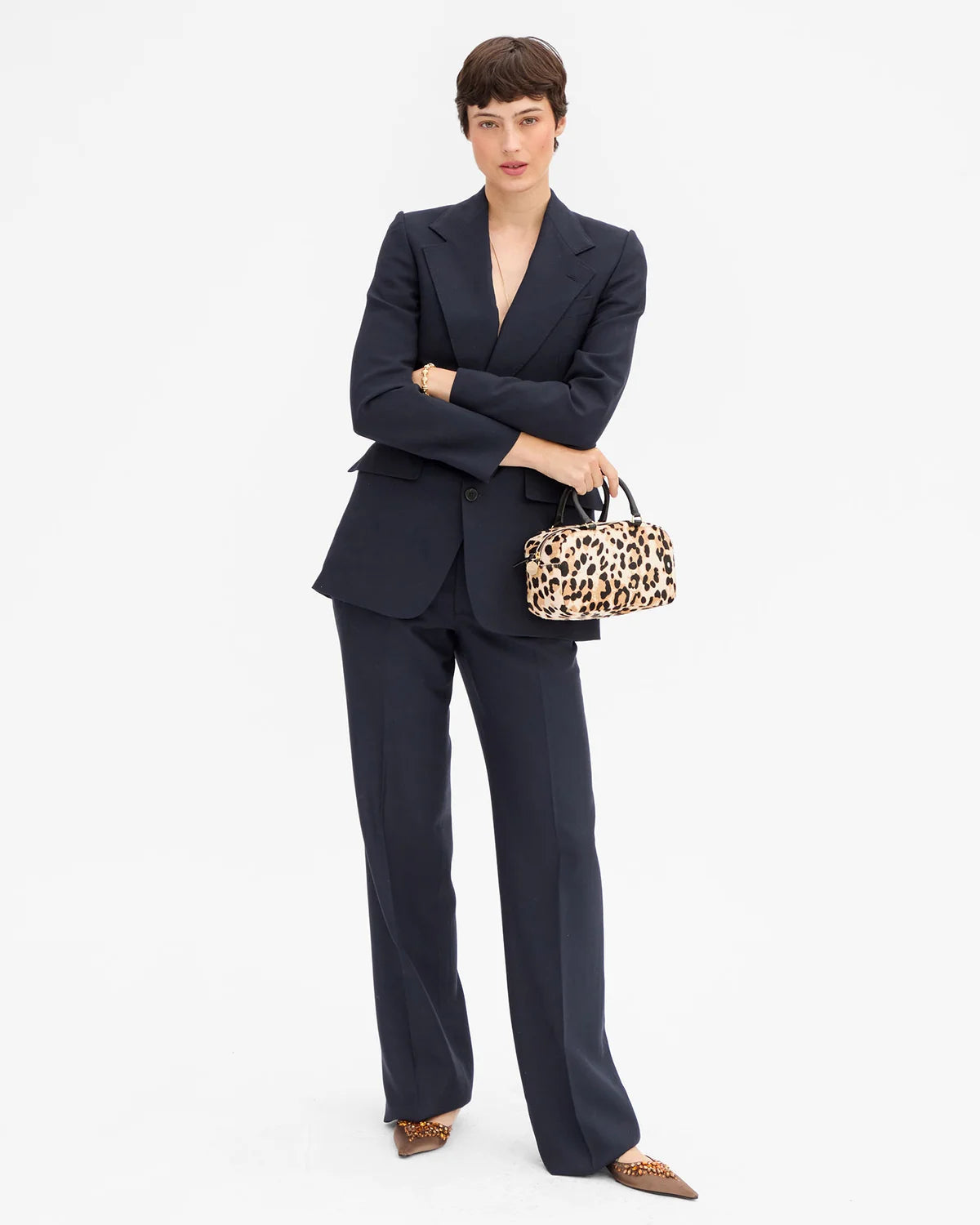 A model stands against a plain white studio background wearing a tailored navy suit with a V-neck blazer and wide-leg trousers, holding a leopard-print handbag, and wearing pointed shoes.