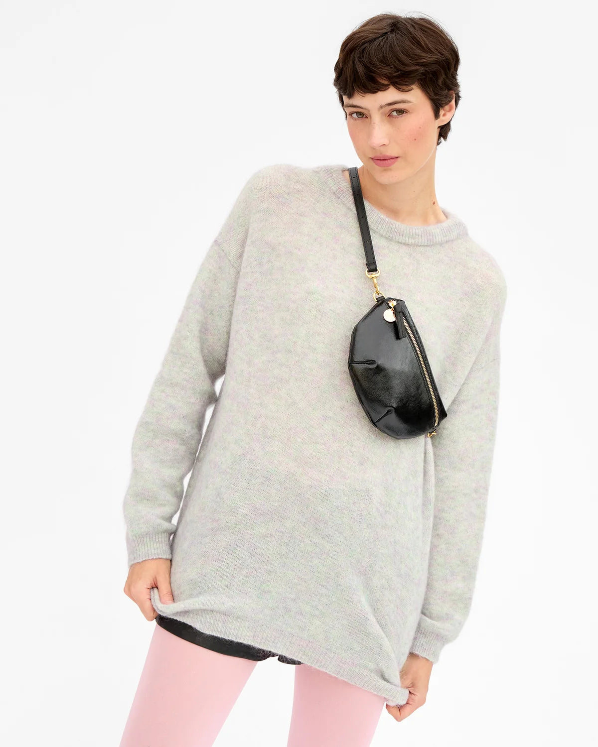 A model wearing an oversized light gray knit sweater, pink leggings, and a black crossbody bag with gold hardware, photographed against a clean white studio background.