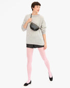 A model wearing a light gray crewneck sweater, black leather shorts, light pink tights, and black shoes, carrying a black faux-leather crossbody bag with gold hardware against a plain white background.