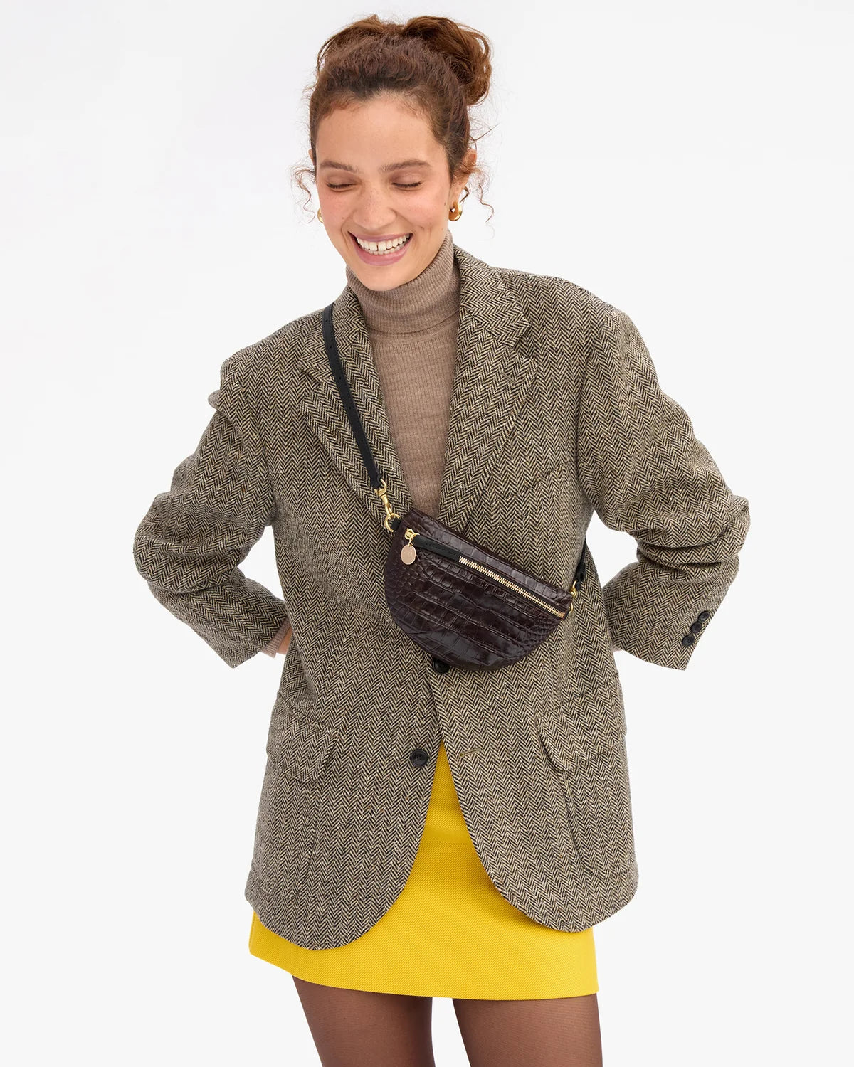 A woman wearing a brown tweed blazer over a brown turtleneck and a yellow skirt, carrying a dark brown crocodile-embossed crossbody bag with gold hardware against a plain white background.