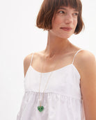 Woman wearing a white top and a necklace with a green heart pendant on a plain background
