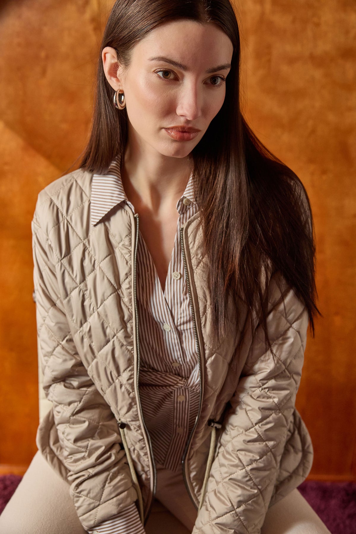 A woman wearing a beige quilted zip-up jacket over a striped shirt, posed against a textured orange-brown background.