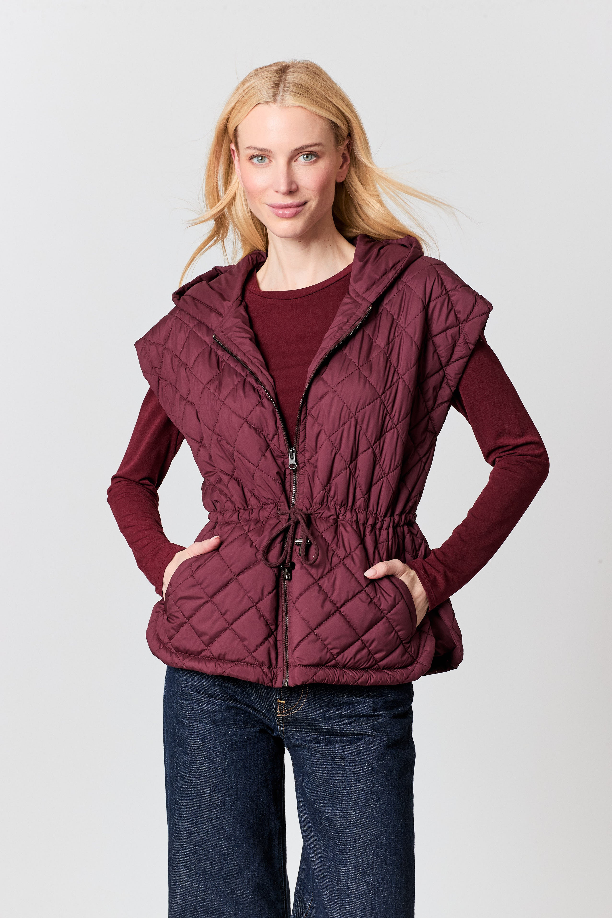 A woman modeling a sleeveless quilted hooded vest in deep burgundy, worn over a matching long-sleeve top and dark blue jeans, against a light gray studio background.