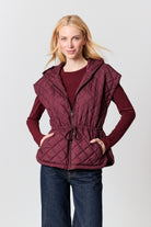 A woman modeling a sleeveless quilted hooded vest in deep burgundy, worn over a matching long-sleeve top and dark blue jeans, against a light gray studio background.