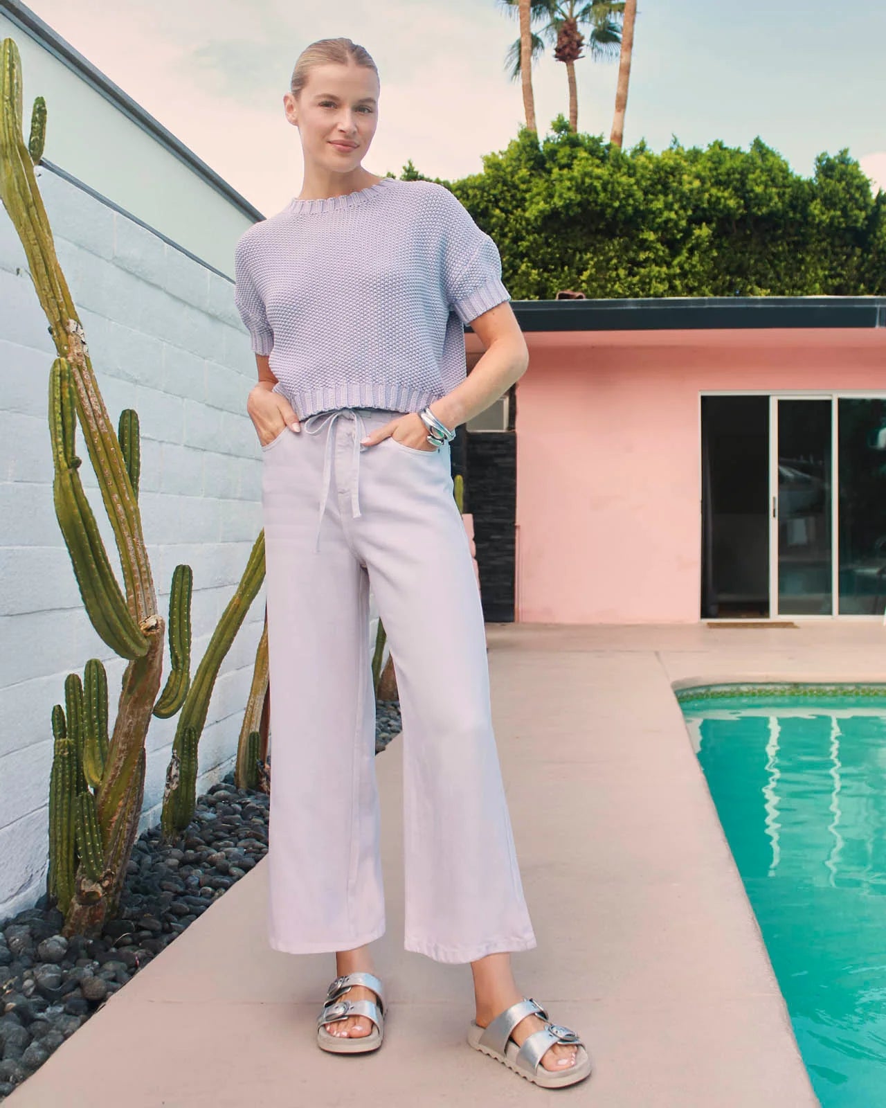 Person standing by a pool with cacti and a pink building in the background