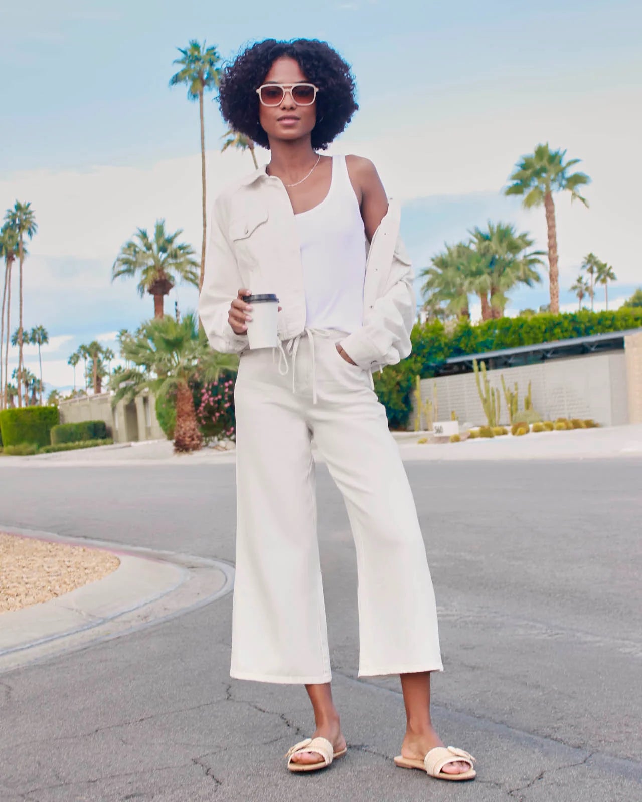 Woman in a white outfit holding a coffee cup with palm trees in the background