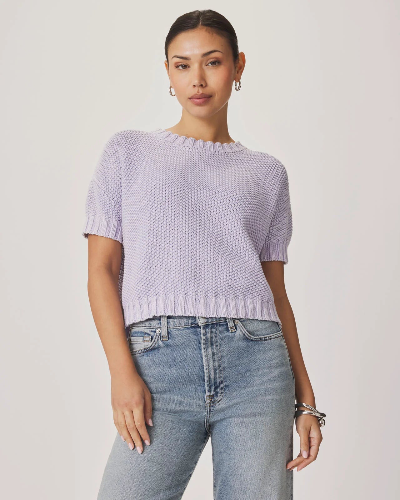 Woman wearing a light purple knitted top and blue jeans on a plain background