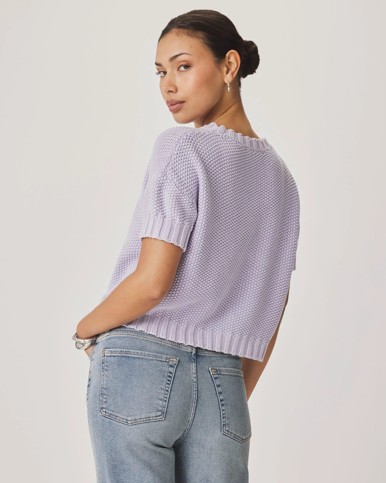 Woman wearing a light purple knitted top and blue jeans on a plain background
